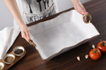 Person wearing a KITCHEN apron holds a white ceramic serving tray with wavy gold trim and gold ring handles on a dark wooden table with spice bowls, red tomatoes, and garlic