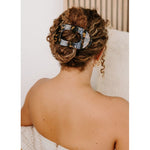 Back of a woman with curly brown hair styled in a low bun, secured by a large oval tortoise-shell hair clip, showcasing bare shoulders and delicate gold hoop earrings against a soft, light background
