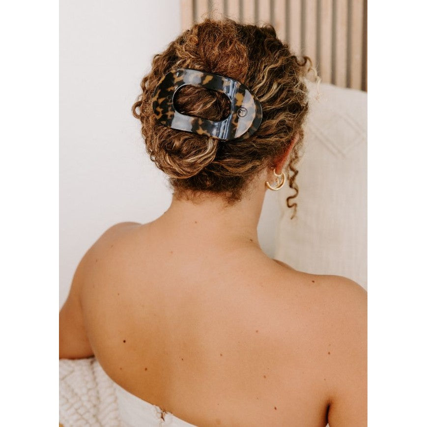 Back of a woman with curly brown hair styled in a low bun, secured by a large oval tortoise-shell hair clip, showcasing bare shoulders and delicate gold hoop earrings against a soft, light background