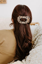 The back of a woman's head indoors shows long auburn wavy hair secured with an oval cream and black leopard print hair clip while wearing a khaki top