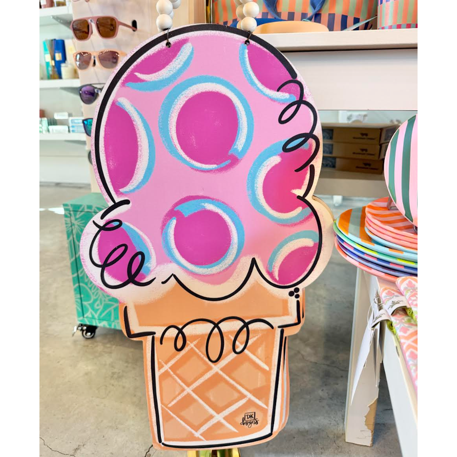 Painted wood sign by DK designs featuring a large pink ice cream scoop with magenta and light blue polka dots, black swirly accents, and an orange waffle cone, displayed in a retail store next to colorful striped plates