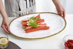 A person wearing a striped KITCHEN apron holds a white oval platter with a gold beaded rim, presenting pink and yellow carrot sticks garnished with frilly green lettuce, on a white table with a glass of lemon water and a bowl of cherry tomatoes