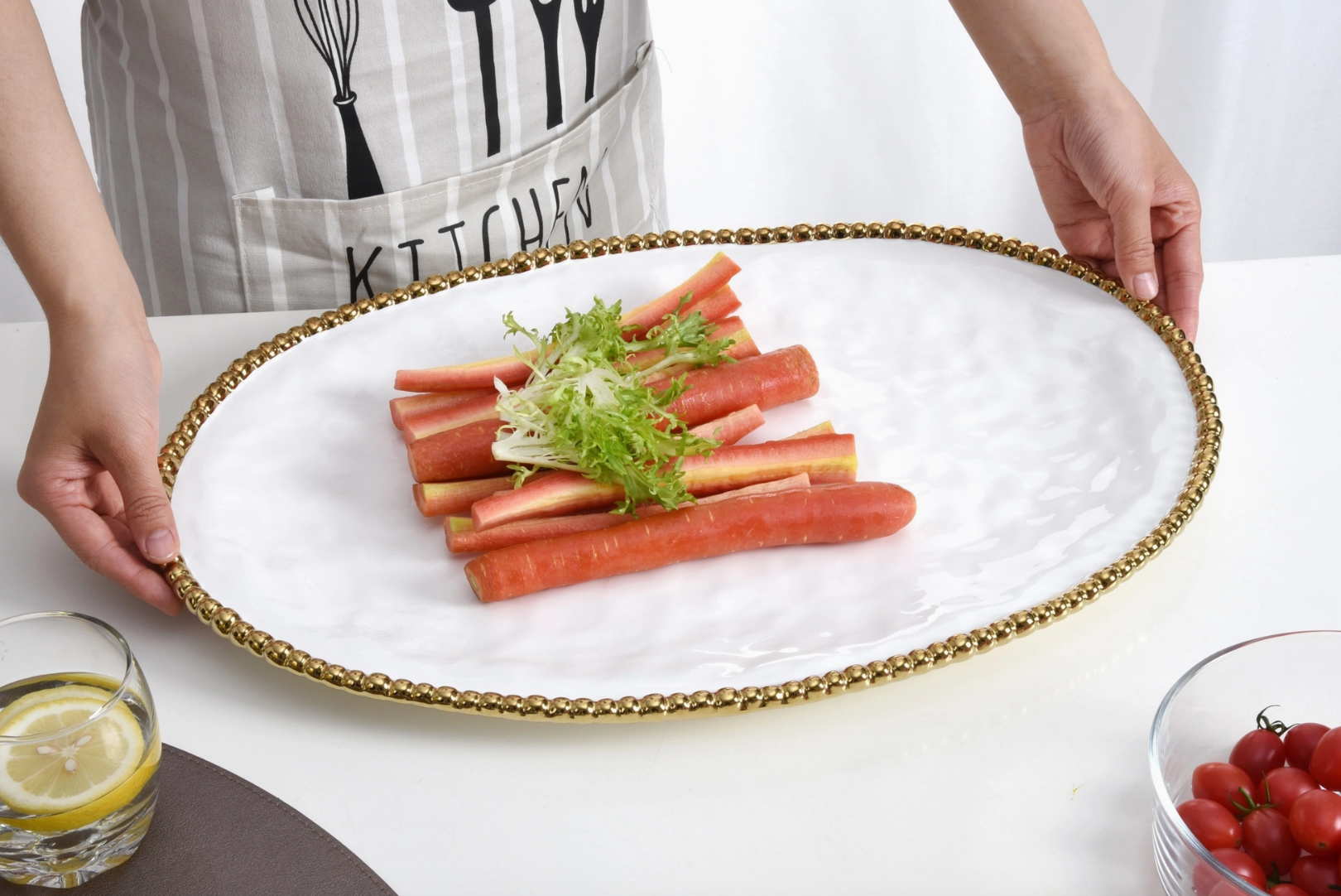 A person wearing a striped KITCHEN apron holds a white oval platter with a gold beaded rim, presenting pink and yellow carrot sticks garnished with frilly green lettuce, on a white table with a glass of lemon water and a bowl of cherry tomatoes