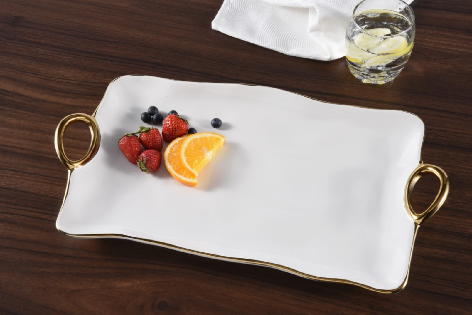 Photograph of a white wavy serving tray with gold handles and trim holding vibrant red strawberries deep blue blueberries and bright orange slices on a dark wooden table next to a glass of lemon water and a white textured napkin