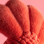 Photograph of a plush coral-orange object featuring three fuzzy ribbed segments and a gathered base set against a soft pink backdrop