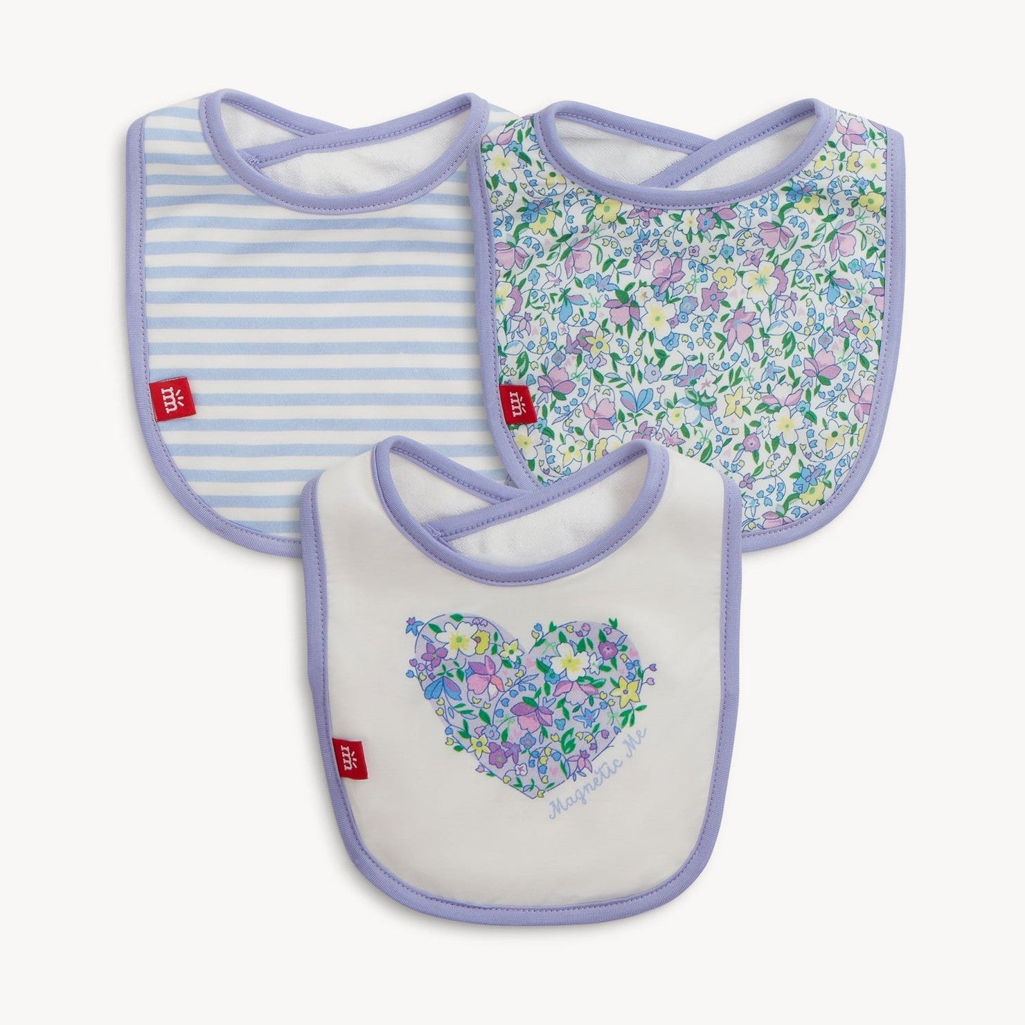 Product photography of three baby bibs including one with blue and white stripes, another with a small multicolored floral pattern, and a white bib featuring a floral heart design with Magnetic Me text, all edged with lilac trim and a red m logo tag