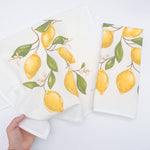 Textile print by Keke M, two white kitchen towels displaying a watercolor-style pattern of bright yellow lemons, vibrant green leaves, and delicate white blossoms on thin brown branches, with one towel unfolded and another neatly folded