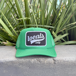 Vibrant green trucker hat with a black embroidered patch featuring white "locals only" text in script and small font, resting on a textured concrete ledge with lush green spiky plants in the background