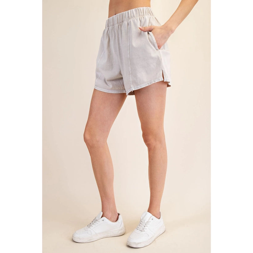 Person modeling light gray elastic waist shorts with a side slit and a hand in the pocket, paired with white lace-up sneakers featuring a subtle pink lining against a warm beige studio background