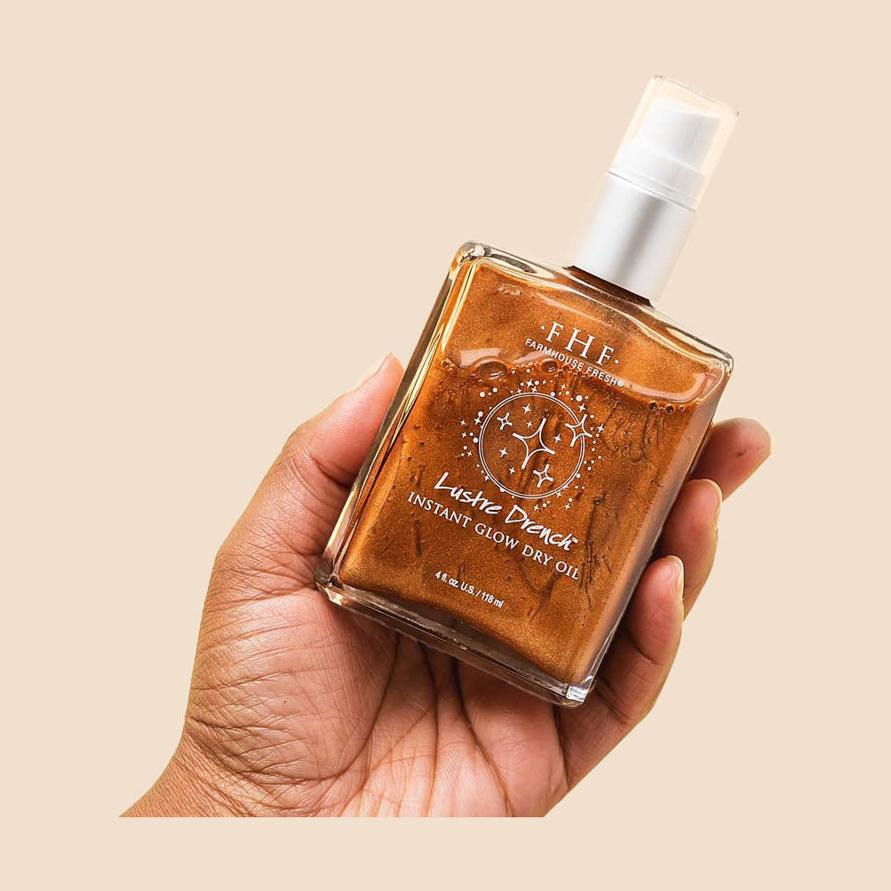 Product photograph of a dark-skinned hand holding a clear rectangular bottle of Farmhouse Fresh Lustre Drench Instant Glow Dry Oil, featuring a shimmering bronze liquid and white pump dispenser, against a plain peach studio background