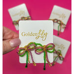 Golden Lily handcrafted beaded bow earrings made with vibrant green, magenta, and gold beads with dangling tails, presented on a white display card against a bright pink background
