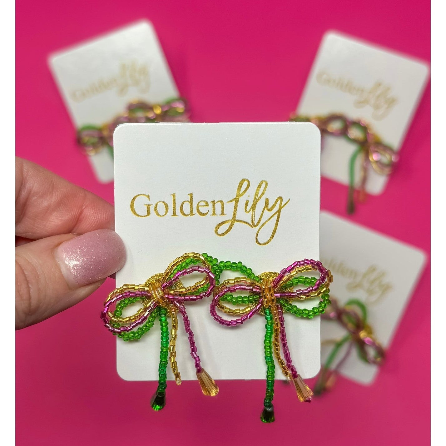 Golden Lily handcrafted beaded bow earrings made with vibrant green, magenta, and gold beads with dangling tails, presented on a white display card against a bright pink background