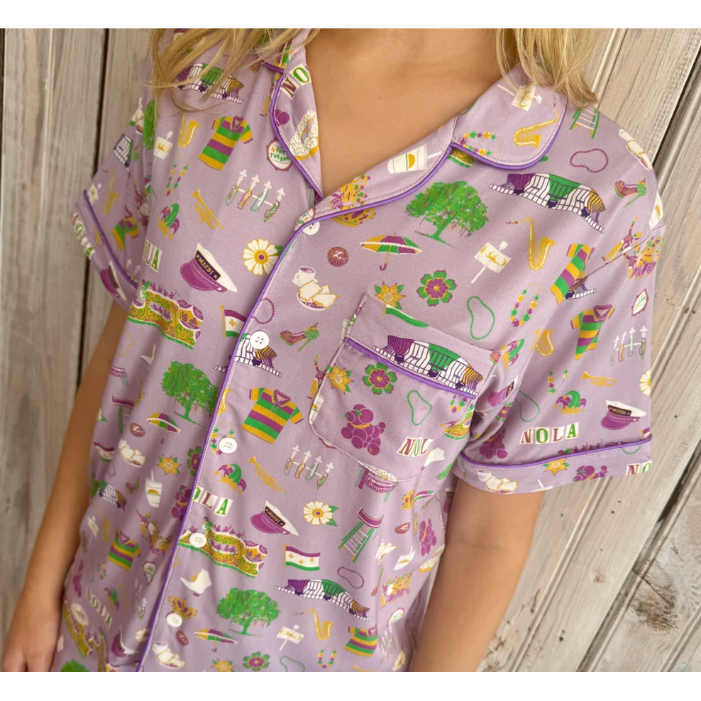 Woman in a light lavender short-sleeved pajama shirt with a New Orleans Mardi Gras pattern featuring the St Charles Streetcar historic balconies jazz instruments and purple green and gold beads