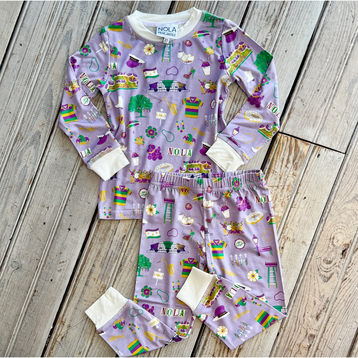 NOLA MERCANTILE children's pajama set depicting New Orleans culture and landmarks including the Superdome, Bourbon Street sign, Mardi Gras masks, and beignets in vibrant purple, green, and gold