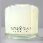 MAGNOLA MAGAZINE branded candle in a clear glass jar filled with white opaque wax featuring a black logo with a stylized fleur-de-lis on a light grey background