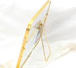 A gold metallic picture frame with clear glass and a wire easel stand, shown from a side perspective, resting on white lace fabric