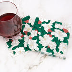 A glass of dark red wine rests next to a stack of decorative paper napkins featuring white magnolia flowers, green leaves, and red berries on a white marble surface