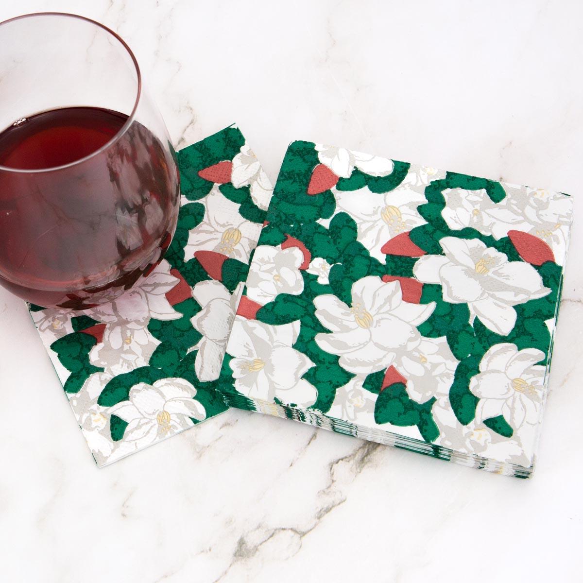A glass of dark red wine rests next to a stack of decorative paper napkins featuring white magnolia flowers, green leaves, and red berries on a white marble surface