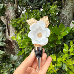 Photograph of a handcrafted wine stopper featuring a white magnolia flower with gold leaves, held by a hand against a vibrant green garden background