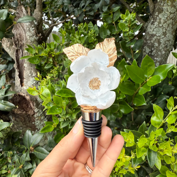 Photograph of a handcrafted wine stopper featuring a white magnolia flower with gold leaves, held by a hand against a vibrant green garden background