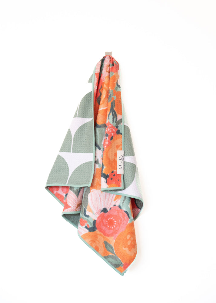 Textile by crae a waffle-knit towel hanging on a white wall featuring a vibrant orange and pink floral pattern on one side and a sage green geometric design on the other