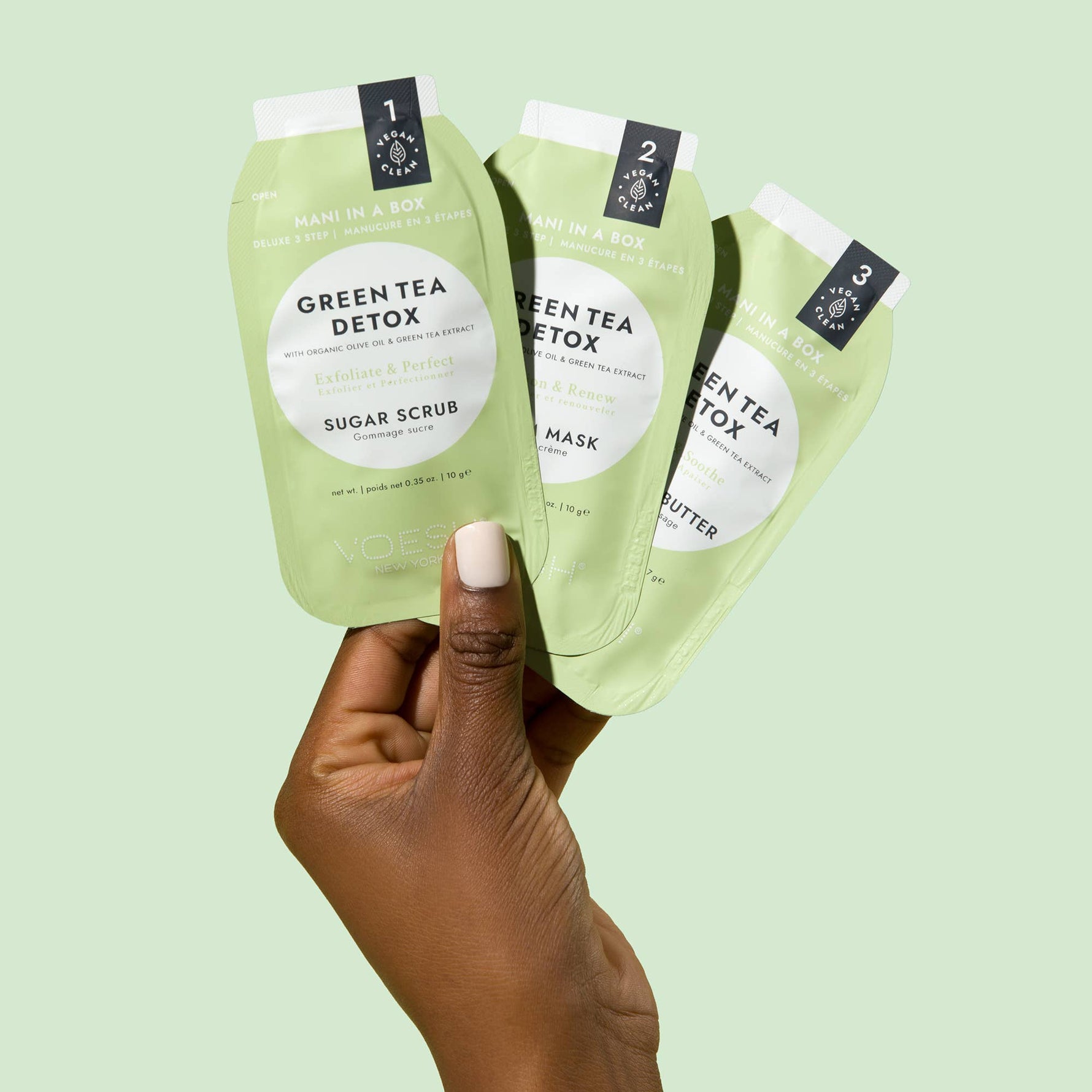 A dark-skinned hand with a light pink manicured thumb holds three light green Voesh New York Mani in a Box Green Tea Detox packets numbered 1, 2, 3, clearly showing the Vegan Clean logo