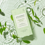 Product photo of a VOESH Mani Moments Green Tea Detox DIY manicure kit floating on a clear rippled water surface surrounded by fresh green leaves and displaying a "Vegan Clean" certification