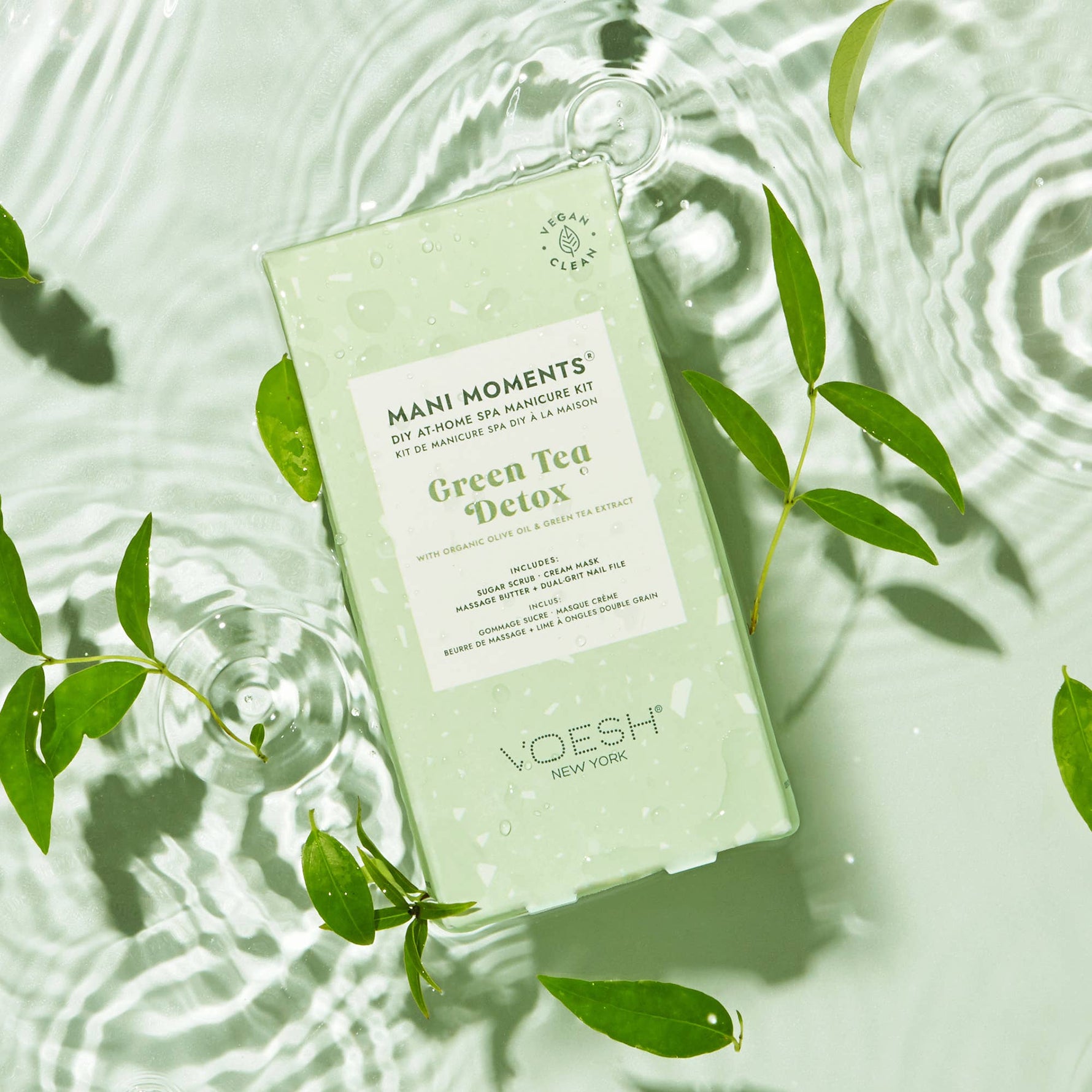 Product photo of a VOESH Mani Moments Green Tea Detox DIY manicure kit floating on a clear rippled water surface surrounded by fresh green leaves and displaying a "Vegan Clean" certification