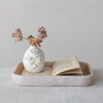 Photograph of a still life on a light tabletop featuring a white ceramic vase with raised floral motifs holding dried pink and brown flowers, placed beside an open vintage book with aged pages, all within a pale pink textured resin tray