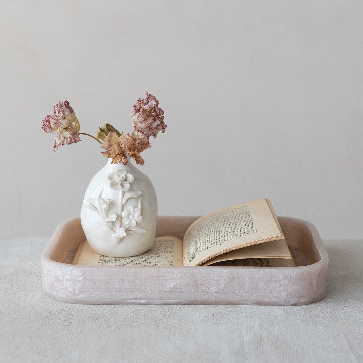 Photograph of a still life on a light tabletop featuring a white ceramic vase with raised floral motifs holding dried pink and brown flowers, placed beside an open vintage book with aged pages, all within a pale pink textured resin tray