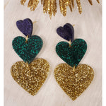 A pair of cascading heart earrings featuring sparkling glitter finishes in royal purple, emerald green, and brilliant gold on a light textured surface