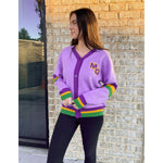 A young woman modeling a lavender cardigan with dark purple trim, a gold MG monogram, and green and yellow striped Mardi Gras themed cuffs and hem, standing outside against a light brick wall and reflective window