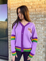 A woman models a vibrant purple V-neck cardigan featuring yellow MG initials on the chest and colorful Mardi Gras-themed purple yellow and green stripes on the sleeves and hem, posed against a textured brick wall outdoors