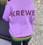 Back of a person in a lilac purple knit sweater with gold and purple KREWE lettering and Mardi Gras purple gold and green striped hem and cuffs against a light brick wall