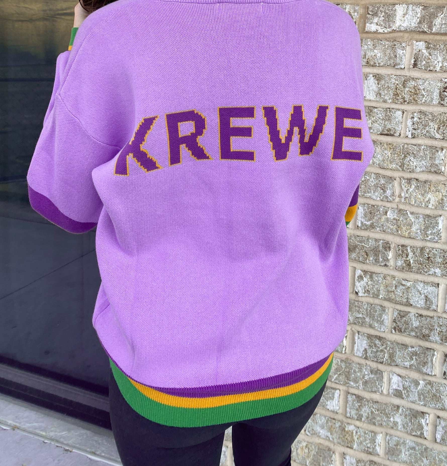 Back of a person in a lilac purple knit sweater with gold and purple KREWE lettering and Mardi Gras purple gold and green striped hem and cuffs against a light brick wall