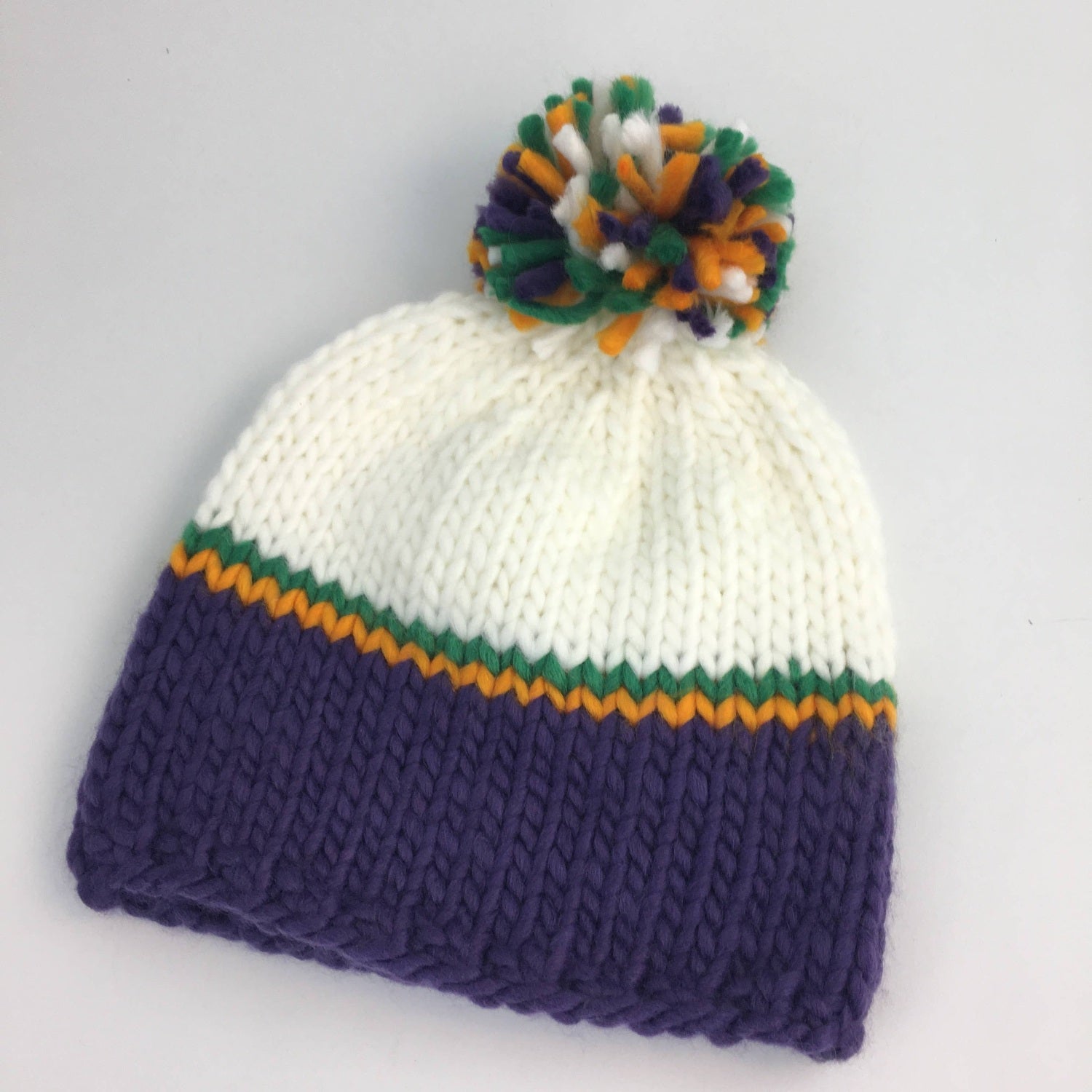 Hand-knitted hat, a Mardi Gras-themed winter beanie with a white crown, purple ribbed base, thin green and gold stripes, and a vibrant multicolored pom-pom