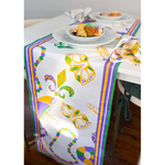 Mardi Gras themed table runner on a dining table showcasing purple, green, and gold masks, fleur-de-lis, beads, and umbrellas, with a slice of King Cake