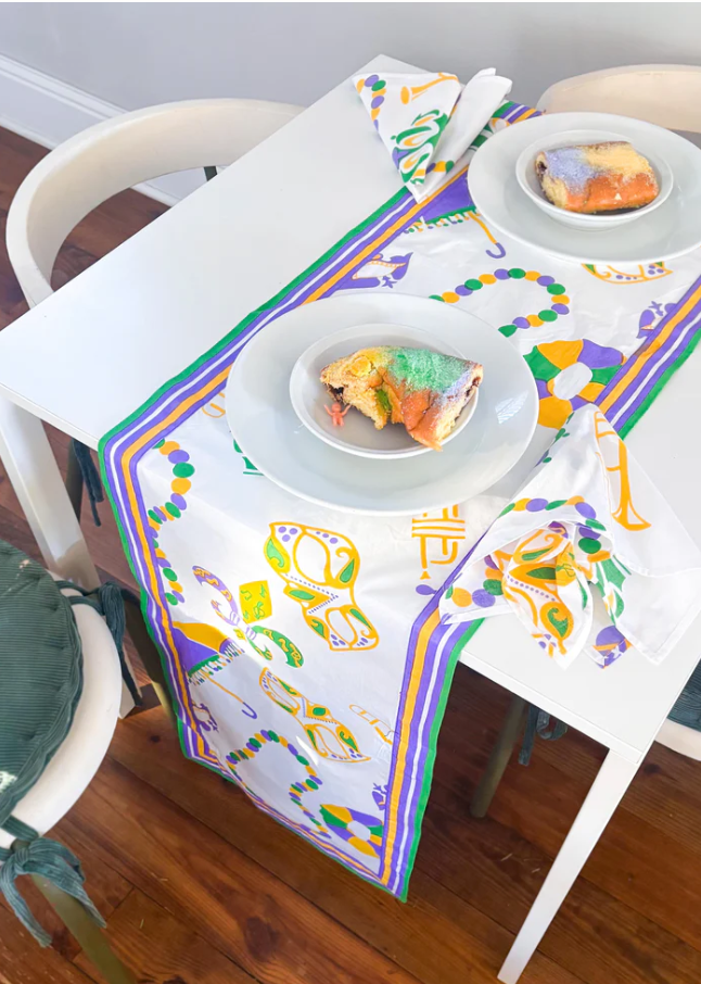A festive Mardi Gras table setting on a white table featuring King Cake slices with traditional purple green and gold icing and a tiny baby figurine alongside a matching patterned table runner and napkins with masks beads and trumpets