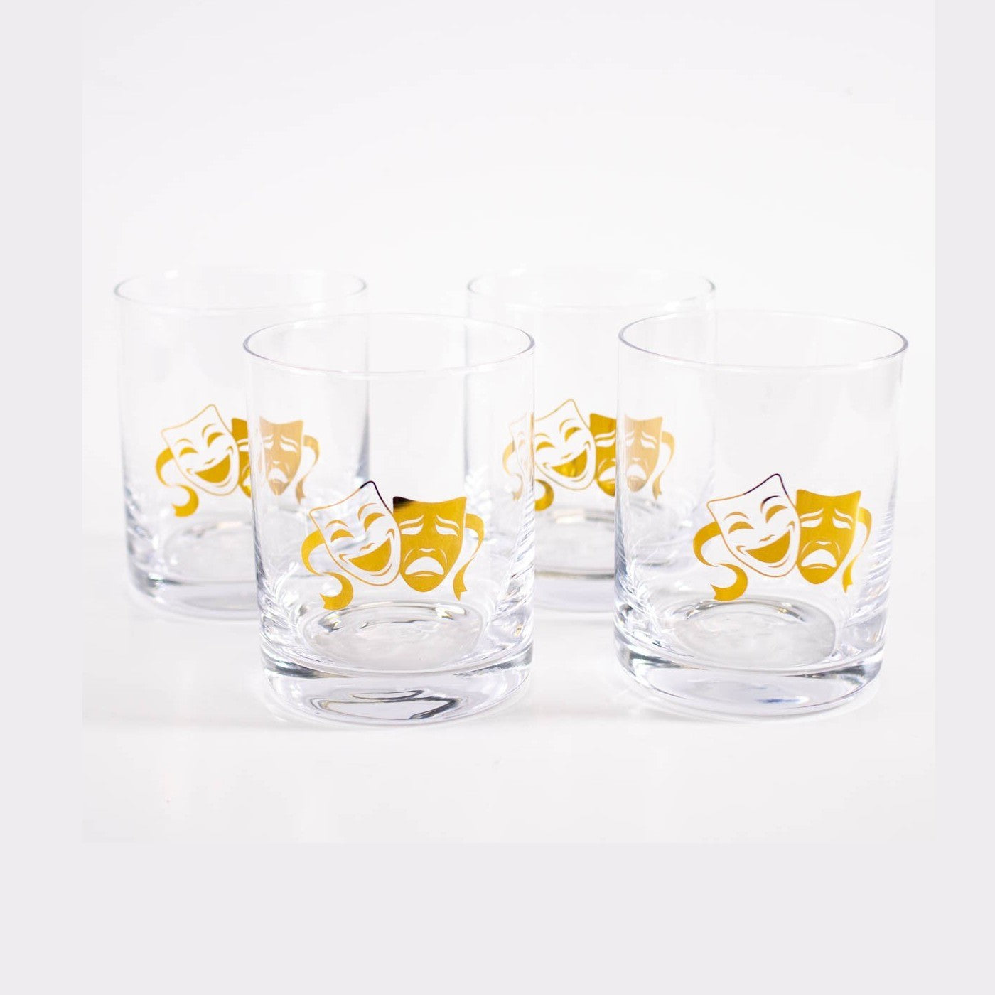 Glassware A set of four clear glass tumblers each featuring a vibrant gold theatrical comedy and tragedy mask design on a bright white background