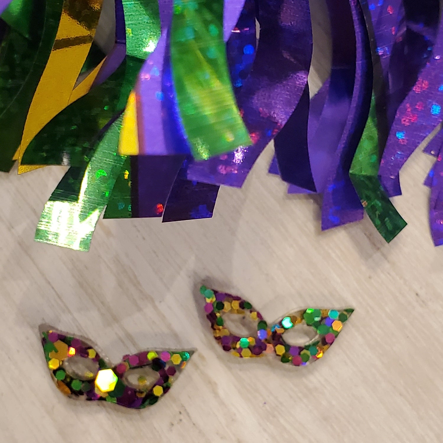 Photograph of festive Mardi Gras decorations with shimmering green, gold, and purple holographic tinsel streamers above two small glitter-covered masks featuring hexagonal glitter pieces