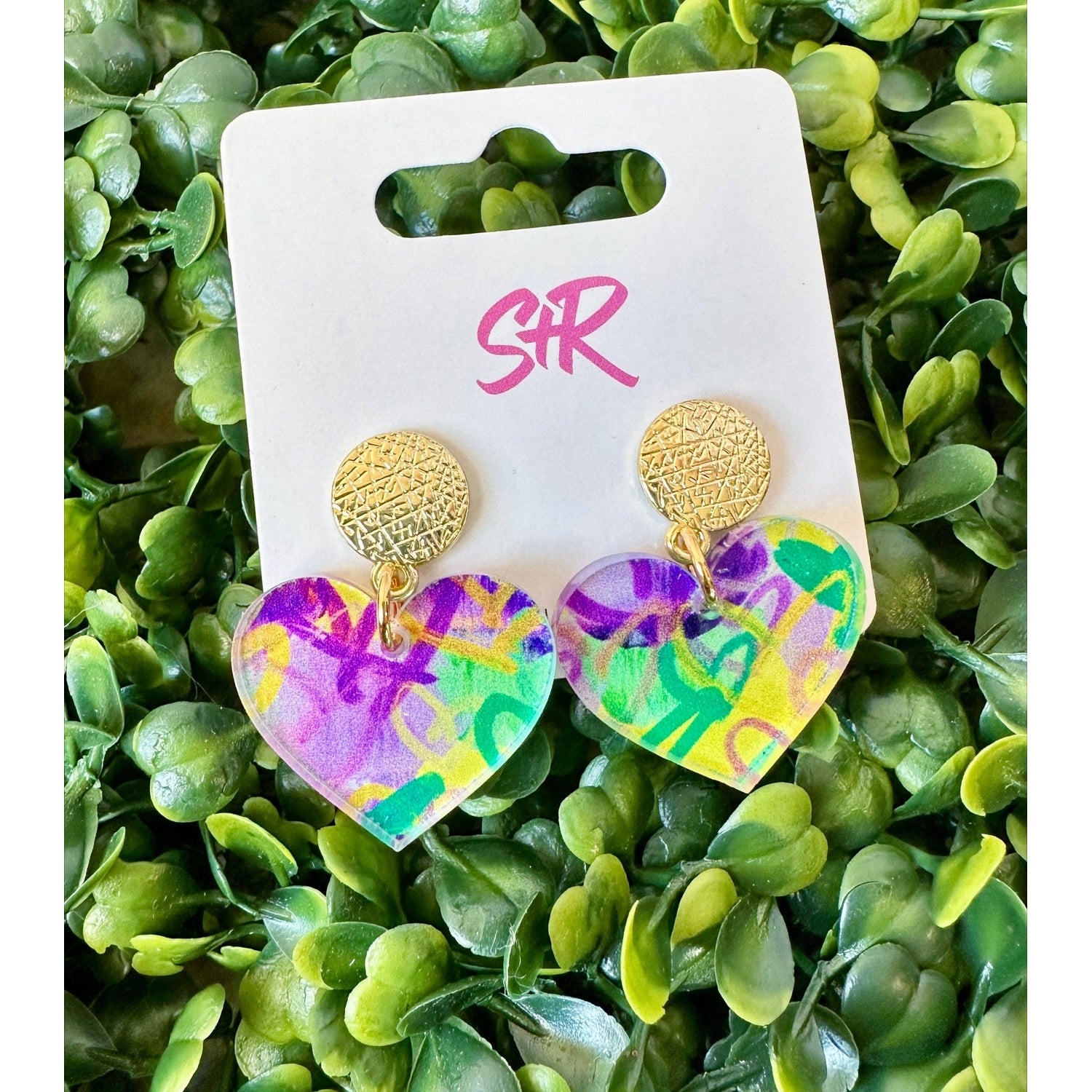 SR jewelry features a pair of acrylic heart-shaped earrings with colorful abstract purple, yellow, and green swirls, topped with textured gold studs and displayed on lush green artificial foliage
