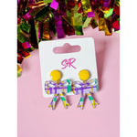 A pair of S+R brand bow-shaped earrings featuring yellow circular studs and translucent acrylic with a purple, green, and yellow plaid pattern displayed on a white card against a pink background with shimmering multi-colored tinsel fringe above