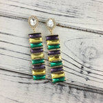 A pair of Mardi Gras-inspired dangle earrings with clear round studs and alternating rectangular emerald green golden yellow and royal purple stones on a distressed white wooden surface