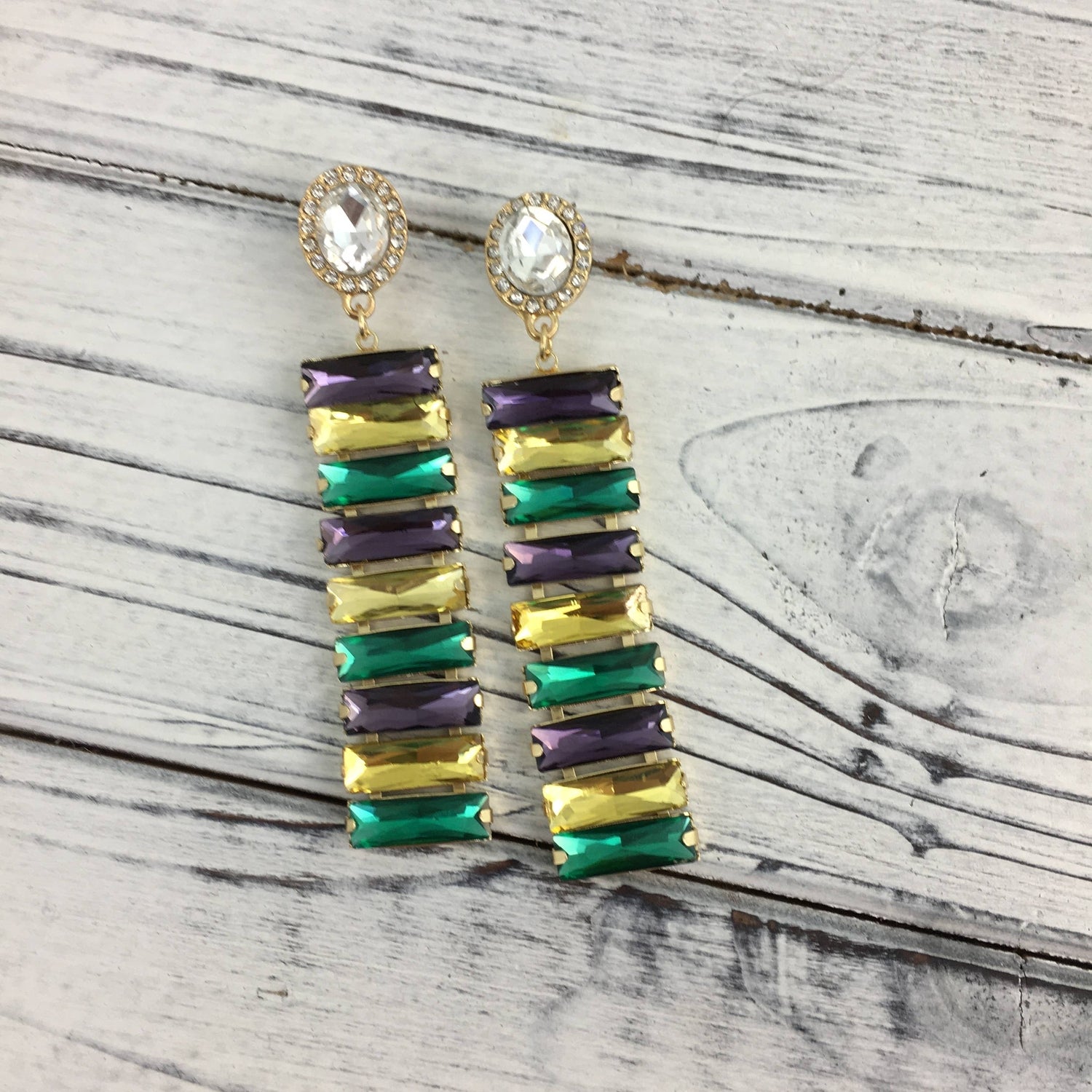 A pair of Mardi Gras-inspired dangle earrings with clear round studs and alternating rectangular emerald green golden yellow and royal purple stones on a distressed white wooden surface