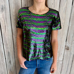 A person modeling a vibrant short-sleeve top covered in iridescent green, purple, and gold sequins paired with blue jeans against a weathered light wood plank wall