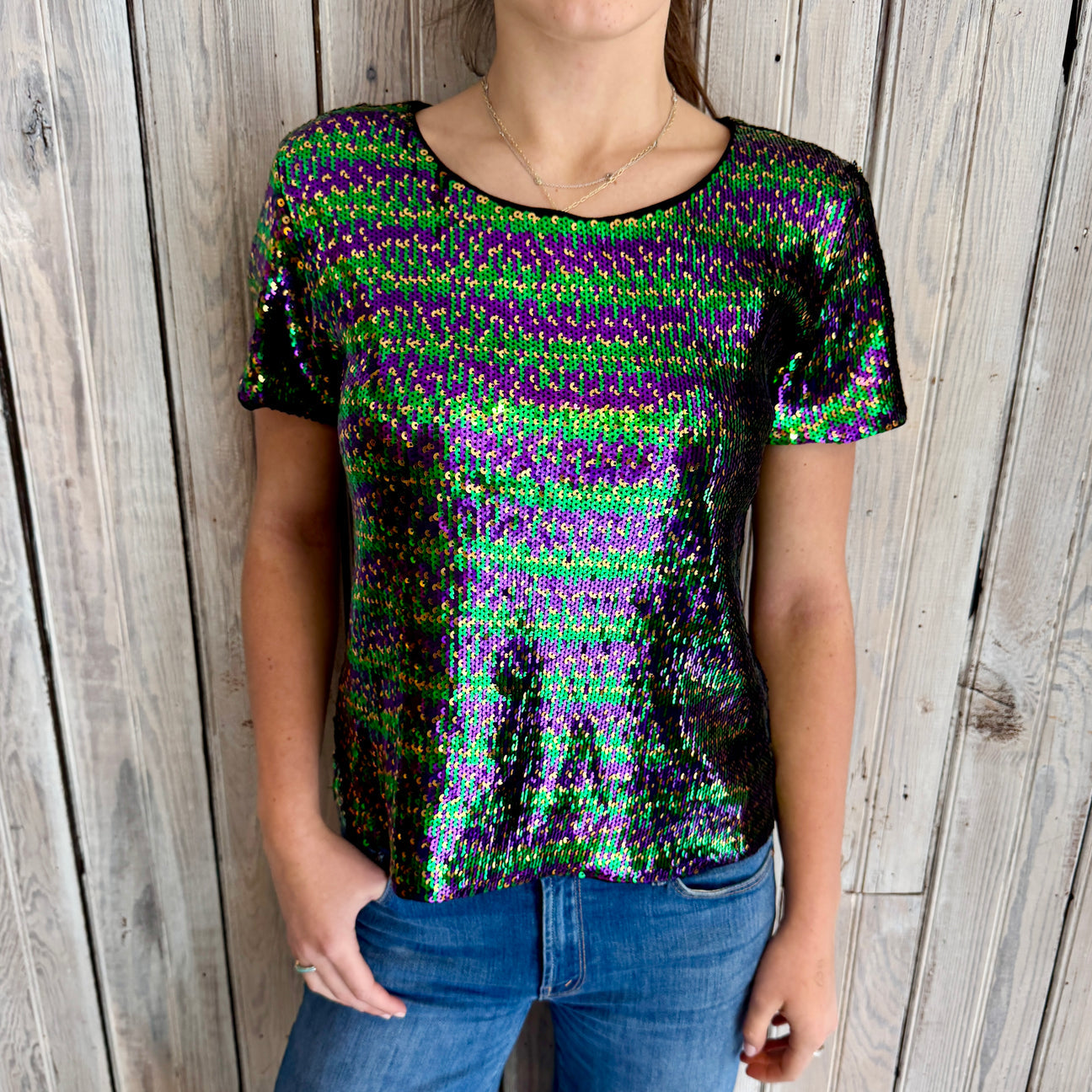 A person modeling a vibrant short-sleeve top covered in iridescent green, purple, and gold sequins paired with blue jeans against a weathered light wood plank wall