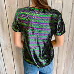 Back of a woman wearing a short-sleeved sequined top shimmering in green, purple, and gold-black against a rustic wooden wall