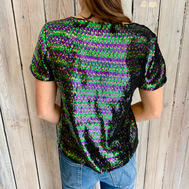 Back of a woman wearing a short-sleeved sequined top shimmering in green, purple, and gold-black against a rustic wooden wall