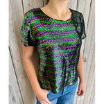 A person models a shimmering short-sleeve top entirely covered in iridescent green, purple, and black sequins, paired with light wash blue jeans and a delicate silver necklace against a weathered wooden wall background
