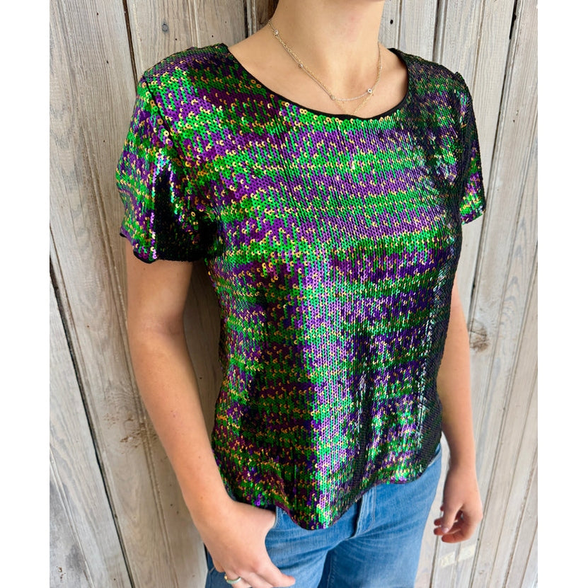 A person models a shimmering short-sleeve top entirely covered in iridescent green, purple, and black sequins, paired with light wash blue jeans and a delicate silver necklace against a weathered wooden wall background